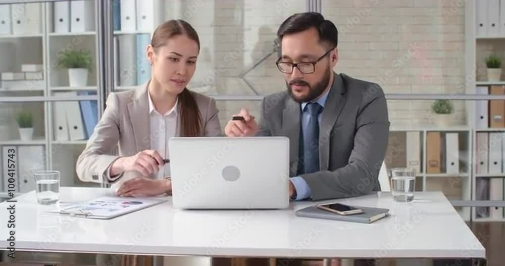 Business partners sitting at office table, looking at laptop display and discussing business report Business partners sitting at office table, looking at laptop display and discussing business report