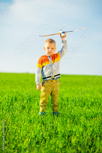 Happy boy playing with toy airplane against blue summer sky and green field background.