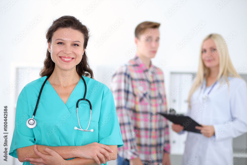 Beautiful young smiling female doctor standing at hospital with doctor and patient on the background. Medical concept. Beautiful young smiling female doctor standing at hospital with doctor and patient on the background. Medical concept.