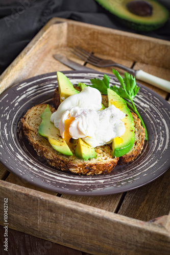 Poached egg and avocado on toasted whole wheat bread with seeds on a plate in vintage wooden tray. Garnished with scallions and parsley.