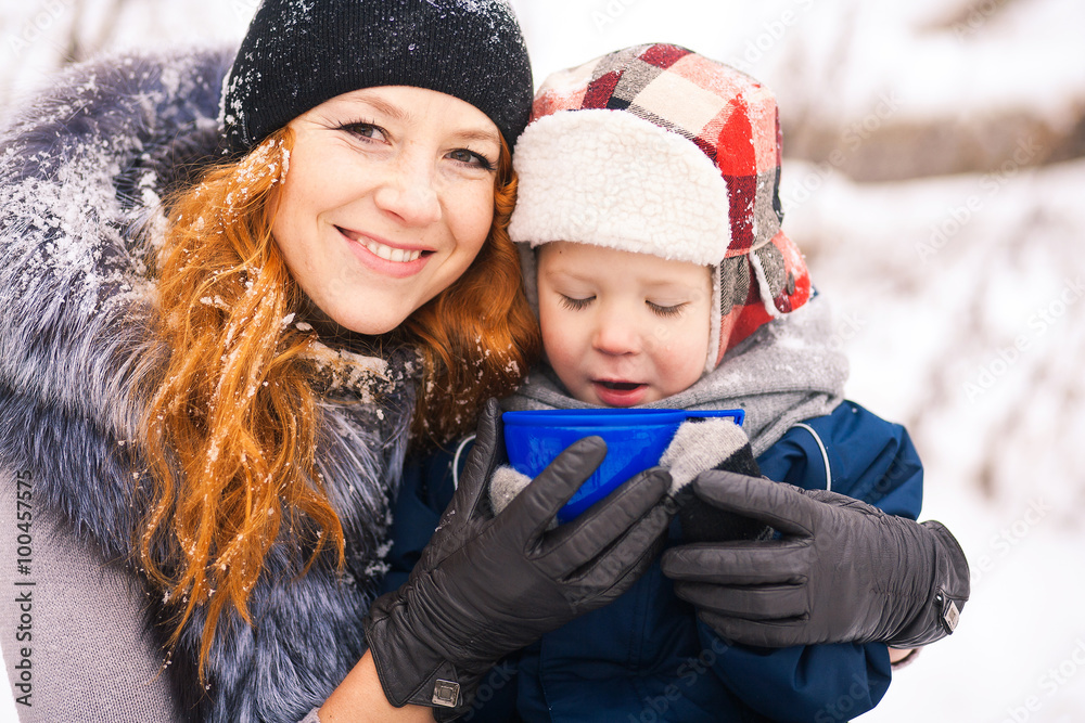 Baby sitting with young beautiful mother outside at snowy trees winter background and drinking hot tee. Happy family enjoying beautiful winter days on picnic.