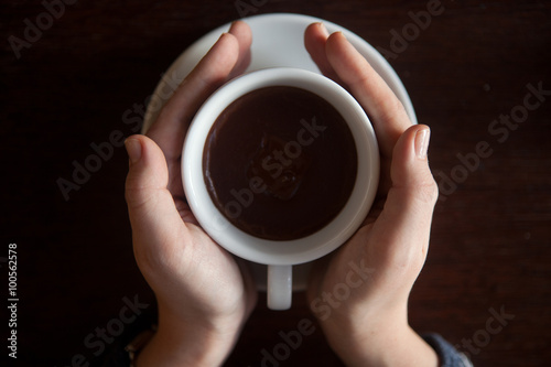 Female hands holding a cup of hot chocolate with foam over wooden table, top view 