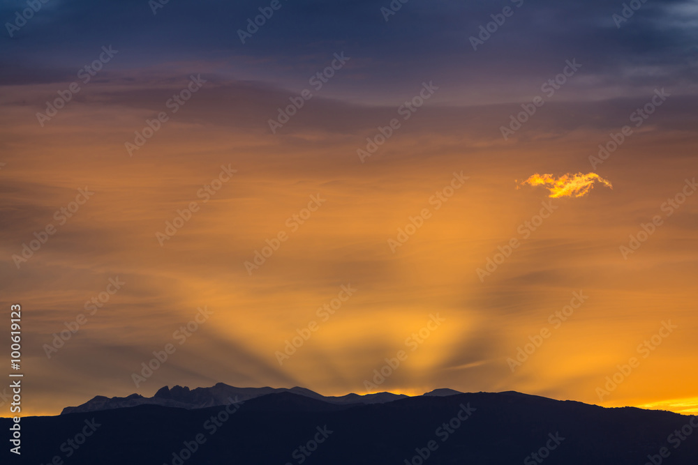 Sun behind dark mountain silhouettes, with colorful sky and clouds