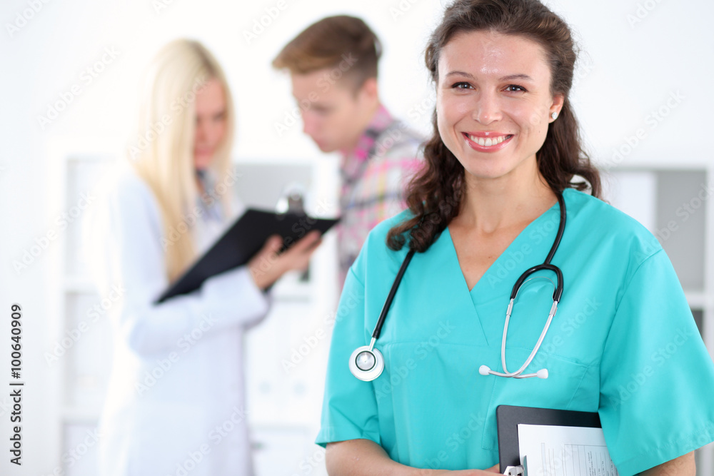 Beautiful young smiling female doctor standing at hospital with doctor and patient on the background. Medical concept. Beautiful young smiling female doctor standing at hospital with doctor and patient on the background. Medical concept.