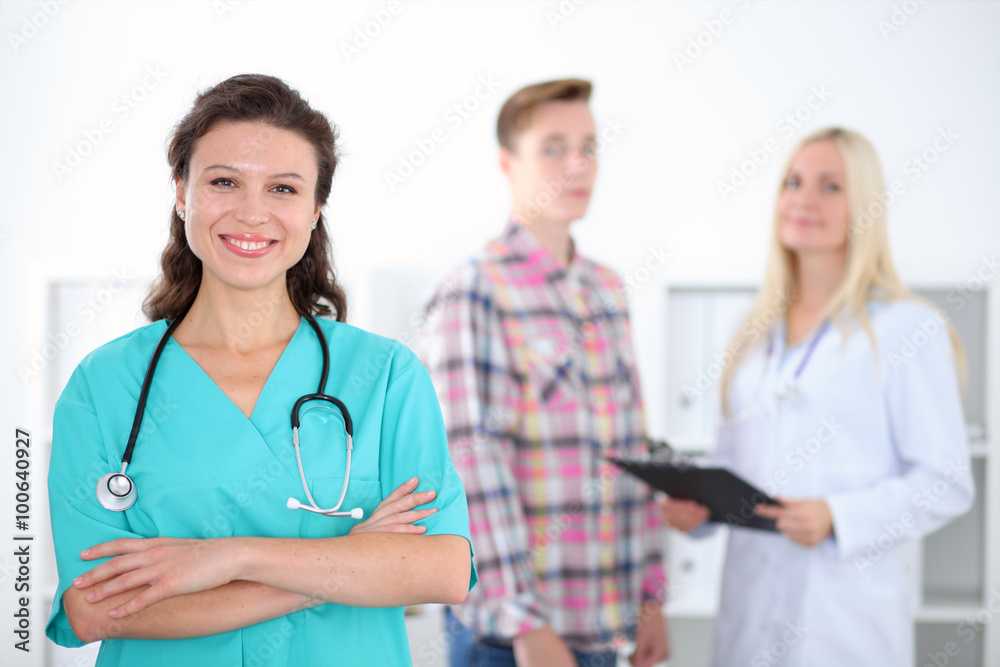 Beautiful young smiling female doctor standing at hospital with doctor and patient on the background. Medical concept. Beautiful young smiling female doctor standing at hospital with doctor and patient on the background. Medical concept.