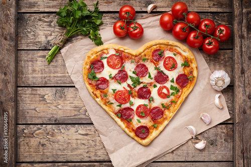 Pizza heart shaped with pepperoni, tomatoes, mozzarella, garlic and parsley on vintage wooden table background. Concept of romantic love for Valentines Day.