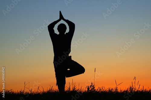 Silhouette of a man practicing yoga in a position Vrikshasana on a grassy horizon after sunset. Silhouette against a bright orange sky.