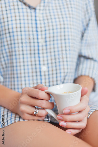 Woman in shirt is sitting on bed with a cup with hot coffee. Bright engagement and wedding rings on her finger.