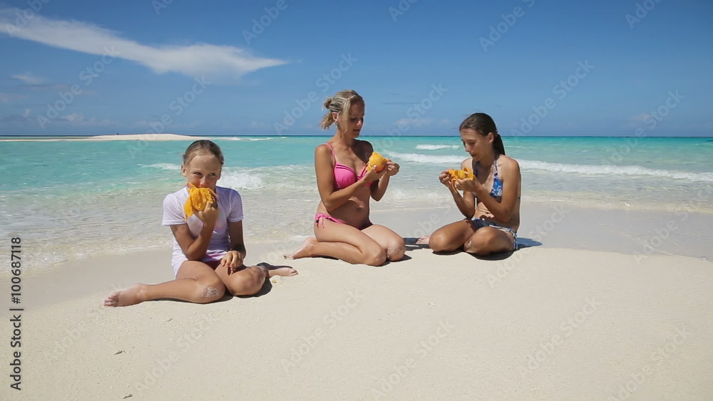Mother and her daughters sitting on a tropical beach and eat mango.happy family eats mango on the shore of tropical sea on a white beach.Travel concept.Family,summer vacation.Happy family Mother and her daughters sitting on a tropical beach and eat mango.happy family eats mango on the shore of tropical sea on a white beach.Travel concept.Family,summer vacation.Happy family