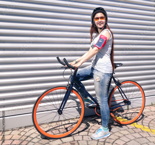  Young happy woman with sport bike and mobile armband standing next to wall - Female cyclist with helmet outdoor in urban area - Concept of alternative transport vehicle in a sunny day