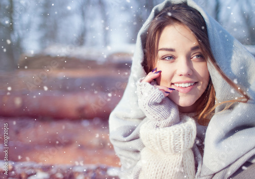 Beautiful smiling happy young woman covered in blanket and snow flakes sitting on pile of pine logs. Snowing winter outdoor leisure concept.