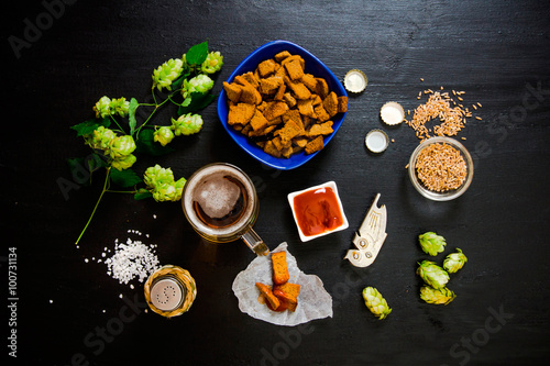 Beer set. Glass of beer, crackers, ketchup, salt. The ingredients for brewing: malt and hops. On a wooden black background.