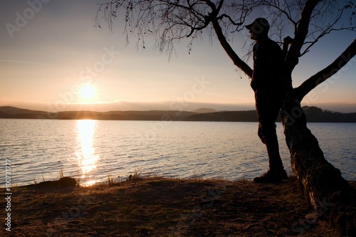 Lone man looking at colorful  sunset on shore of autumn lake. Man stand bellow birch tree. photo