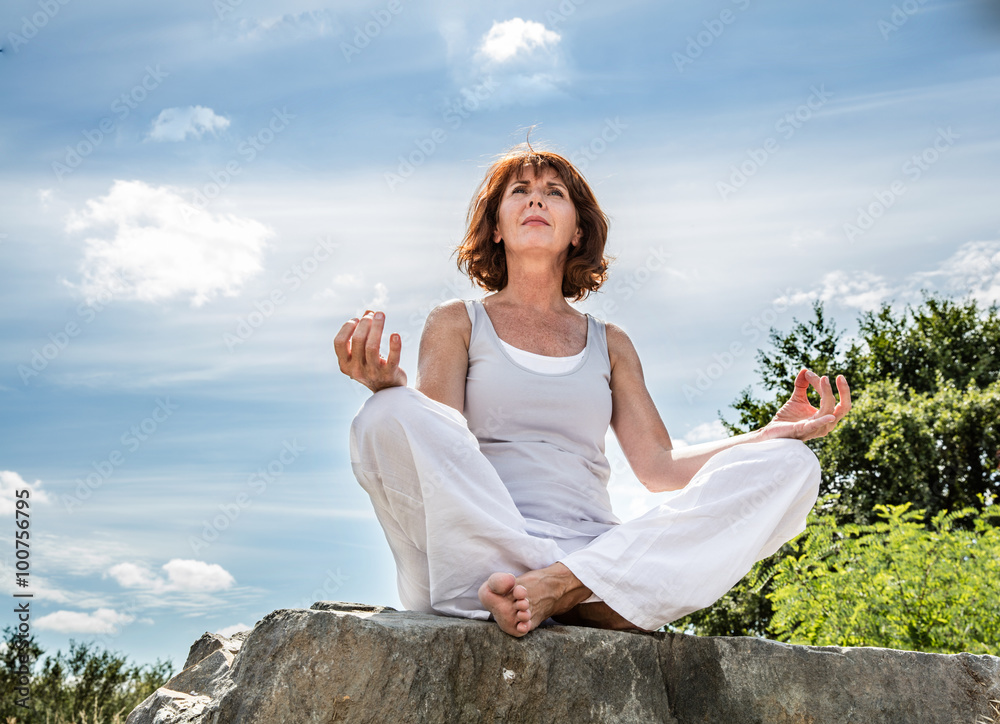 Obraz premium breathing outdoors - beautiful 50s woman sitting on a stone in yoga lotus position, seeking for spirituality over summer blue sky,low angle view