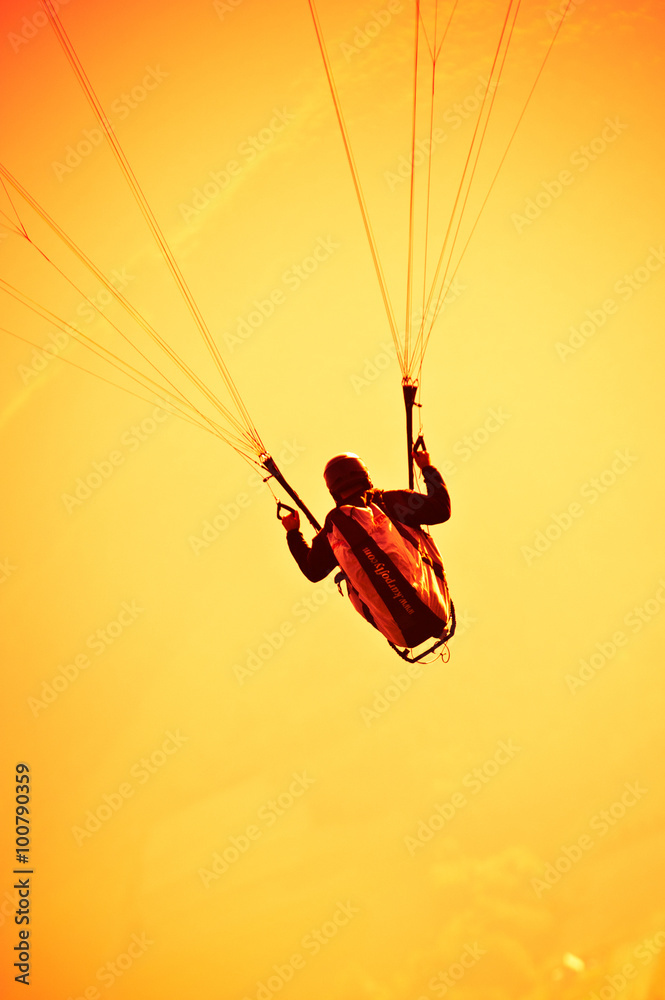 A man flying a paraglider holds steering cables in his hand, orange sky