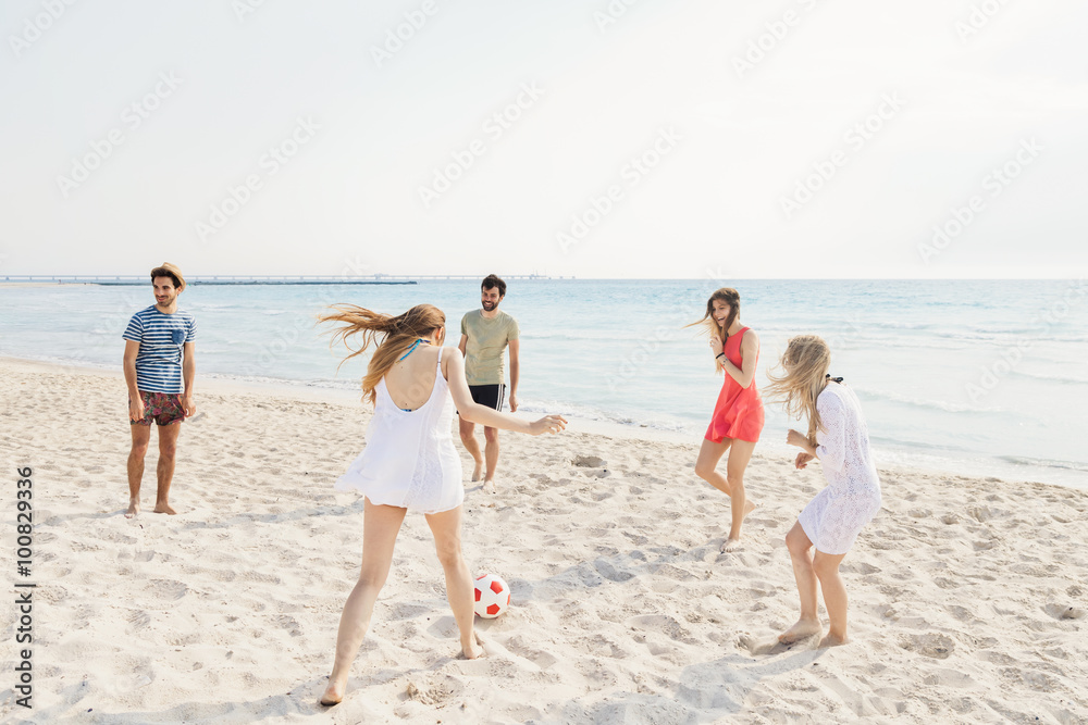 Five friends, three women and two men, playing with a ball of red and white color on the desert sandy beach in a summer day. The beach is fine sand and the sea is turquoise