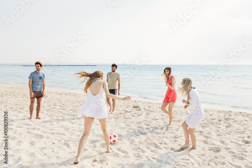 Five friends, three women and two men, playing with a ball of red and white color on the desert sandy beach in a summer day. The beach is fine sand and the sea is turquoise