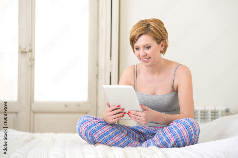 young attractive red hair girl in sleeping tank top and pajamas pants sitting on bed at home using internet digital tablet pad young attractive red hair girl in sleeping tank top and pajamas pants sitting on bed at home using internet digital tablet pad