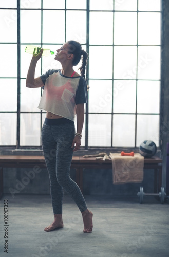 Woman in profile standing in loft gym drinking from water bottle