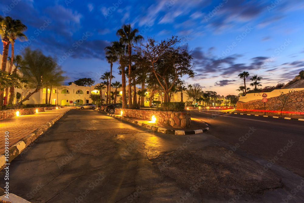 Evening view for road in illumination, white apartments, palm trees Evening view for road in illumination, white apartments, palm trees