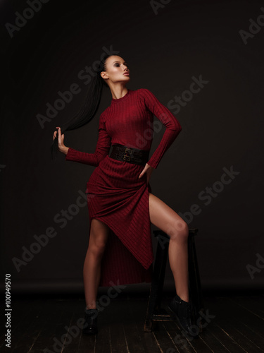 The slim elegant model in the vinous dress and black patent leather boots with slicked straight ponytail and long beautiful legs is posing in the fashion studio