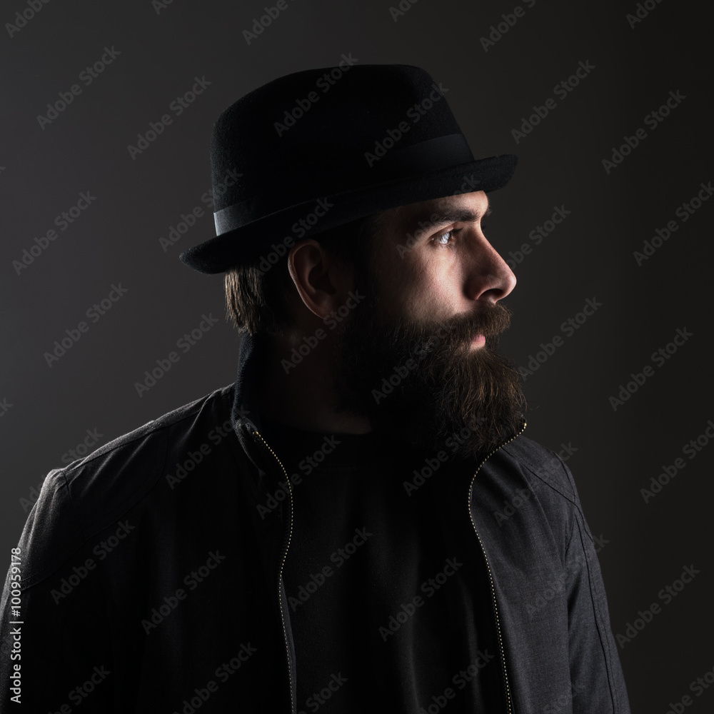 Profile view of bearded man wearing hat looking away. High contrast low key dark shadow portrait over black background. Profile view of bearded man wearing hat looking away. High contrast low key dark shadow portrait over black background.