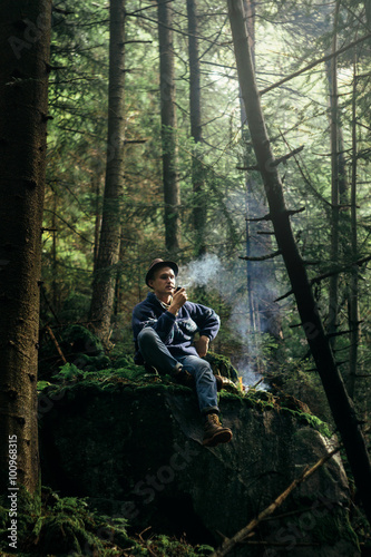 stylish hipster traveler smoking tobacco pipe in sunny forest in