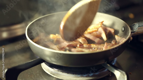 Small pieces of pancetta frying in the skilet and stirring with wooden spoon on gas stove in preparation of spaghetti carbonara photo