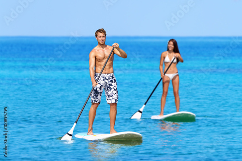 Man and woman stand up paddleboarding on ocean. Young couple are doing watersport on sea. Male and female tourists are in swimwear during summer vacation. photo