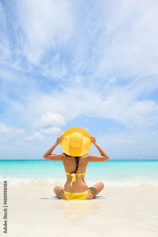 Beach Travel Vacation Holidays Woman Relaxing. Woman sitting on sea shore during summer. Rear view of female wearing yellow sunhat and bikini. Carefree tourist is enjoying vacation at beach. Beach Travel Vacation Holidays Woman Relaxing. Woman sitting on sea shore during summer. Rear view of female wearing yellow sunhat and bikini. Carefree tourist is enjoying vacation at beach.