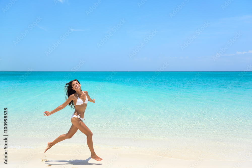 Carefree young woman with arms outstretched jumping on sea shore during summer. Full length of exhilarated female in white bikini. Tourist is enjoying vacation during summer. Carefree young woman with arms outstretched jumping on sea shore during summer. Full length of exhilarated female in white bikini. Tourist is enjoying vacation during summer.