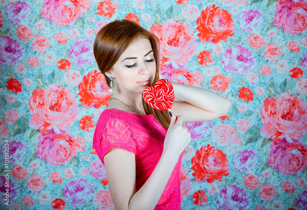 Fototapeta premium Studio portrait of beautiful smiling young woman in a pink dress holding heart shape red lollipop in front of colorful background 