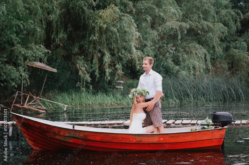 Happy young couple with a bouquet and a wreath hug sitting in a boat on the lake and sky background, lifestyle, love, relationships Happy young couple with a bouquet and a wreath hug sitting in a boat on the lake and sky background, lifestyle, love, relationships