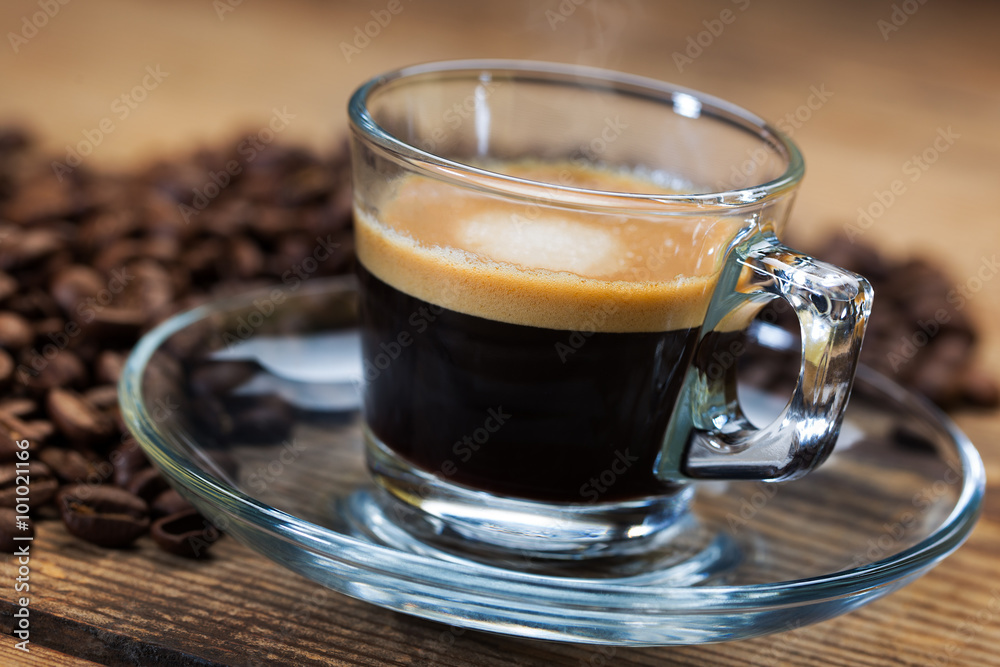 A transparent glass cup of hot steamy espresso on a saucer standing on a wooden table top, surrounded by coffee beans A transparent glass cup of hot steamy espresso on a saucer standing on a wooden table top, surrounded by coffee beans