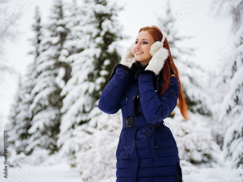 Pretty girl with bright red hair and soft fluffy warm mittens on her hands is wandering around in the white winter forest surrounded by the snow fir-trees