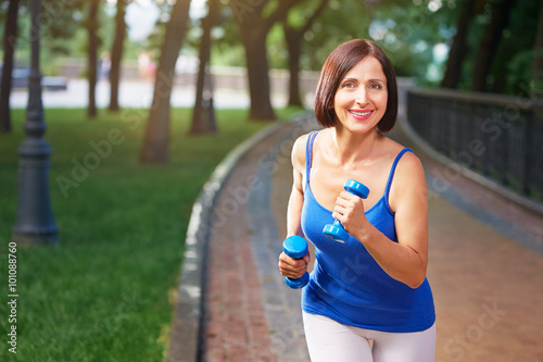 Active senior woman running smiling in the park with bar bells i