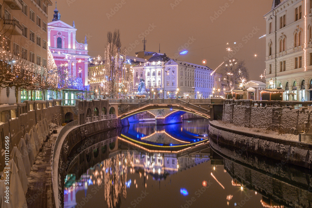 Ljubljana, Slovenia - January 3, 2016. Snowy banks of Ljubljanica river, Triple bridge, on the background with illuminated Franciscan Church of the Annunciation and christmas tree.