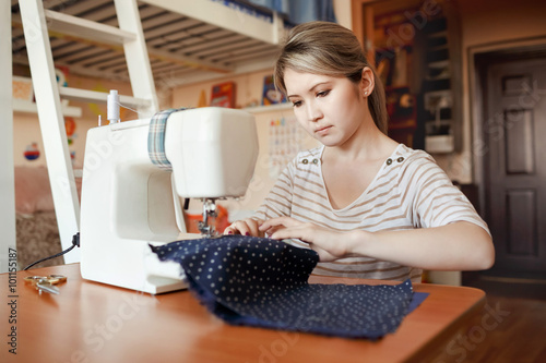 Obraz na plátně Young woman sewing with sew machine at home while sitting by her working place