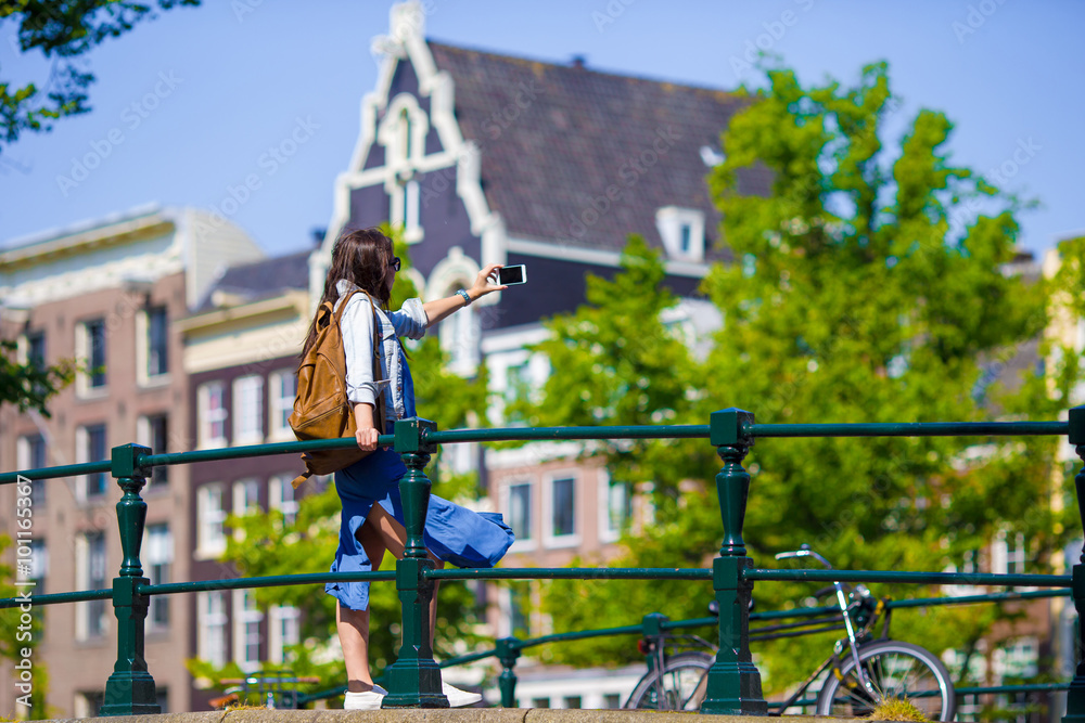 Obraz premium Young woman taking self portrait in european city outdoors. Young adult holding smartphone camera to take a picture of herself during her summer vacation in Amsterdam