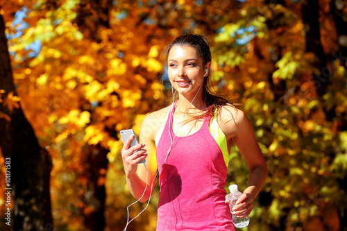Young beautiful woman with bottle of water running in autumn park and listening to music with headphones.