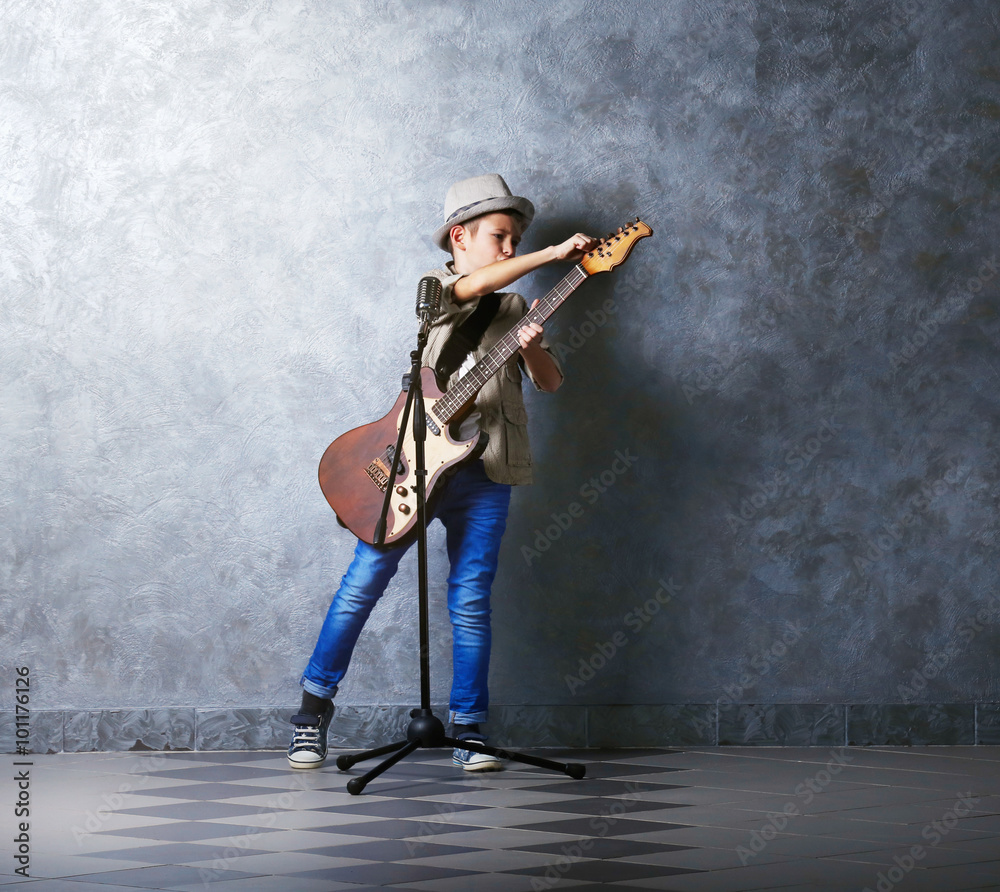 Little boy playing guitar and singing with microphone on a grey wall background Little boy playing guitar and singing with microphone on a grey wall background