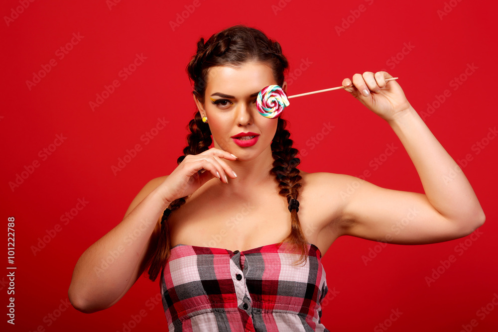 Obraz premium Beauty fashion model girl eating colorful lollipop. Surprised funny young woman with braids hairstyle, nails and makeup beige, isolated on a red background