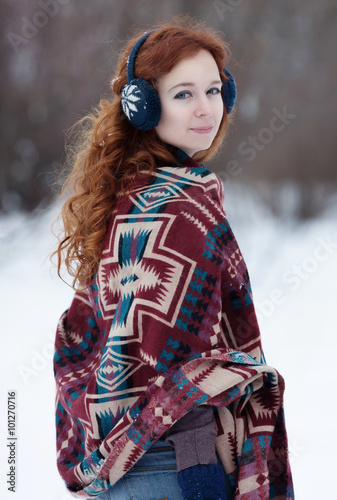 Young beautiful red-haired woman in blue headphones and gloves. She is wrappening in a big scarf with ethnic pattern. She looks over her right shoulder. There is the snowy forest around her.