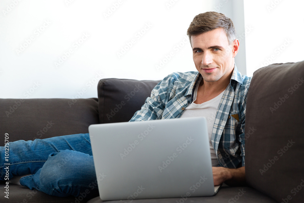 Attractive smiling man sitting on sofa and using laptop Attractive smiling man sitting on sofa and using laptop