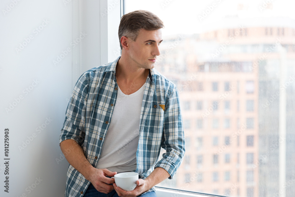 Fototapeta premium Handsome man sitting on windowsill and drinking coffee