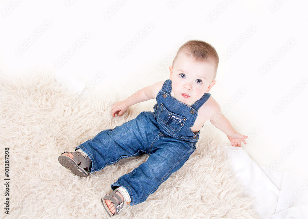 Naklejka premium Cute baby with big blue eyes in denim overalls sitting isolated on white background.