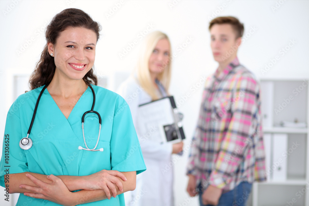 Beautiful young smiling female doctor standing at hospital with doctor and patient on the background. Medical concept. Beautiful young smiling female doctor standing at hospital with doctor and patient on the background. Medical concept.