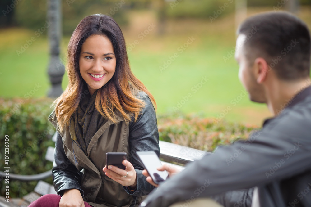 Beautiful young woman using phone and talking to young man while sitting on a bench in a park