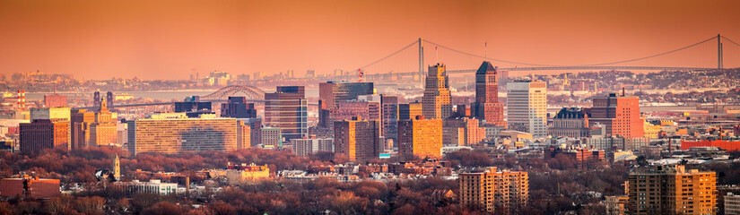 Newark New Jersey skyline viewed from Eagle Rock reservation under an orange sunset. In the background, under a hazy sky, Verrazano bridge links Staten Island to Brooklyn.