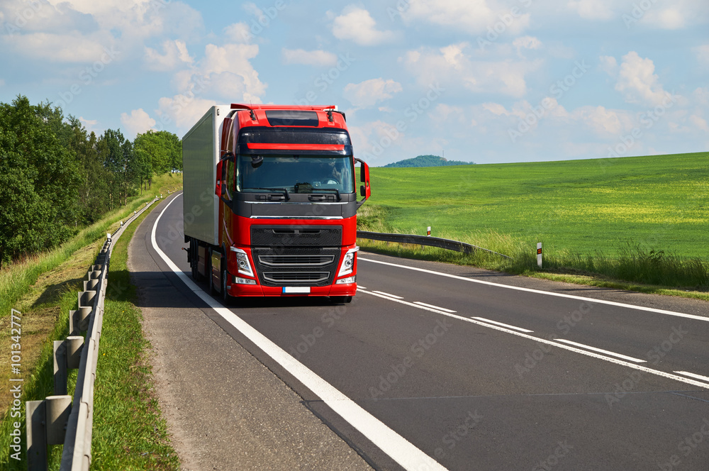 Red truck driving on asphalt road in a rural landscape. Green fields and alleys leading along the road. Sunny summer day with a cloudy sky.